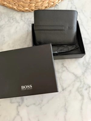 Svart plånbok från BOSS i läder - Stilren svart plånbok från BOSS i äkta läder med präglad logga på framsidan. Plånboken har en klassisk vikbar design och flera fack för kort och sedlar. Levereras i original ask, perfekt för dig som gillar minimalistisk och exklusiv stil. Helt ny. Nypris ca 1200 kr