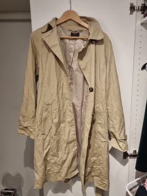 Beige trenchcoat från Jofama - Stilren beige trenchcoat från Jofama med klassisk krage, knäppning framtill och två fickor. Lätt material och helfodrad insida gör den perfekt för lager på lager. Snyggt avslappnad passform och tidlös design.