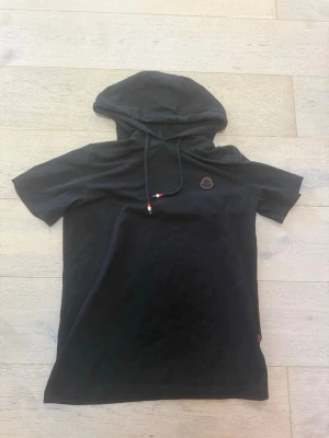 Svart kortärmad t-shirt med huva - Snygg svart T-shirt med korta ärmar och huva med dragsko. Liten broderad detalj på bröstet i rött och vitt. Perfekt för en avslappnad streetwear-look. Tillverkad i mjukt bomullstyg för skön känsla.