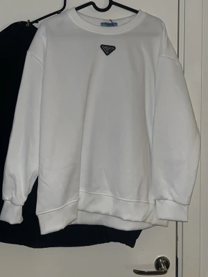 Vit sweatshirt med liten logga - Säljer en clean vit sweatshirt med rund hals och en liten svart logga på bröstet. Tröjan har ribbade muddar vid ärmslut och nederkant. Perfekt för dig som gillar enkel och stilren design.