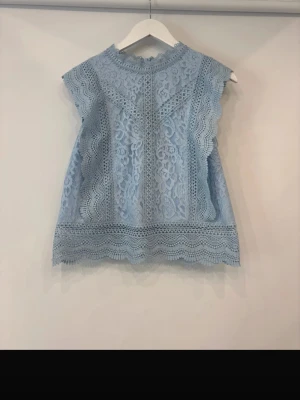 Ljusblå spetsblus med volang - Supersöt ljusblå blus i spets med fina volangdetaljer och hög hals. Ärmlös modell med vackert mönstrad spets och dekorativa kanter. Perfekt för dig som vill ha en romantisk och trendig topp till sommaren.