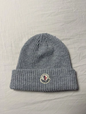 Grå ribbstickad mössa från Moncler - Säljer en stilren grå ribbstickad mössa från Moncler med uppvikt kant och klassisk Moncler-logga framtill. Mössan är gjord i mjukt material och har en enkel, clean look som passar till det mesta. Perfekt för dig som vill ha både värme och stil.