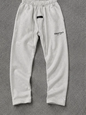 Gråa Essentials mjukisbyxor - Snygga ljusgrå mjukisbyxor från Fear of God Essentials med svart logga på låret och en svart patch framtill. Byxorna har resår i midjan, dragsko och sidofickor. Perfekta för en chill och avslappnad stil.