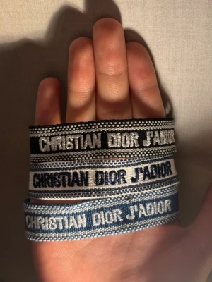 Christian Dior J'Adior tygarmband - Säljer tre snygga tygarmband från Christian Dior med texten 'J'Adior'. Armbanden kommer i svart, grått och blått med kontrasterande broderad text. Perfekt accessoar för att lyfta din outfit och visa din stil. 1 - 159, 2 - 279, 3 - 309
