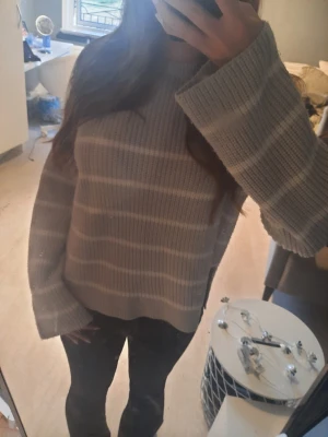 Gråbeige randig stickad tröja - Mysig stickad tröja i gråbeige med breda, ljusa horisontella ränder. Tröjan har en avslappnad passform, långa ärmar med vida muddar och rund halsringning. Perfekt för lager på lager och chill dagar.