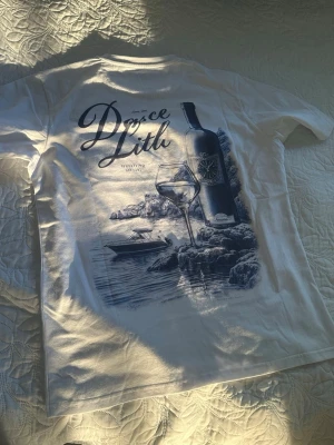 Vit t-shirt med vintema och tryck - Vit t-shirt med stort tryck på ryggen som visar en vinflaska, vinglas och landskap. Texten 'Drøset Litt' syns tydligt. T-shirten har klassisk passform och korta ärmar, perfekt för dig som gillar unika motiv.