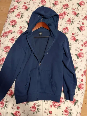 Mörkblå hoodie med dragkedja - En stilren mörkblå hoodie från Fila med dragkedja framtill och två fickor. Tröjan har en klassisk huva och ribbade muddar vid ärmar och nederkant. Perfekt för en avslappnad look och enkel att matcha med jeans eller joggers.