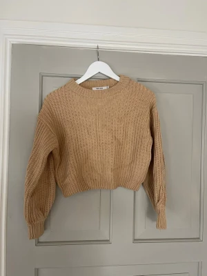 Beige stickad tröja från NA-KD - Säljer en beige stickad tröja från NA-KD i storlek XS. Tröjan har en croppad passform, ribbstickade muddar och rund halsringning. Perfekt för lager på lager och till hösten!