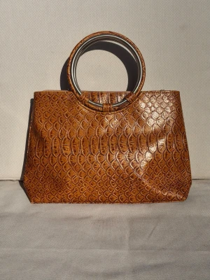 Handväska krokodilmönster - Handväska i en varm, brun nyans med detaljrikt krokodilmönster. Med runda metallhandtag som ger väskan en modern men samtidigt klassisk vintagelook Mycket fint skick Höjd 37 cm (med handtag), bredd 35 cm, djup 8 cm
