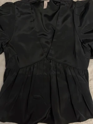 Svart satinblus med puffärm - Stilren svart blus i glansigt satinmaterial med djup v-ringning och puffiga korta ärmar. Blusen har markerad midja och lätt rynkad nederdel för en feminin siluett. Perfekt att styla med jeans eller kjol för en trendig look.