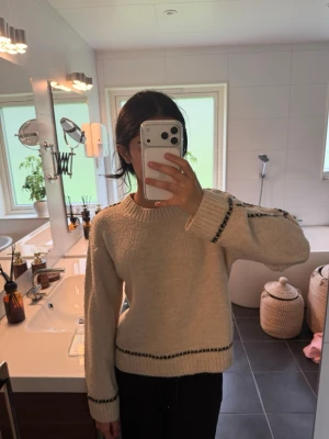Beige stickad tröja med svarta detaljer - Mysig beige stickad tröja med svarta ränder vid ärmslut, nederkant och längs ärmen. Tröjan har en avslappnad passform, rund halsringning och långa ärmar. Perfekt för kalla dagar och enkel att matcha med olika outfits.