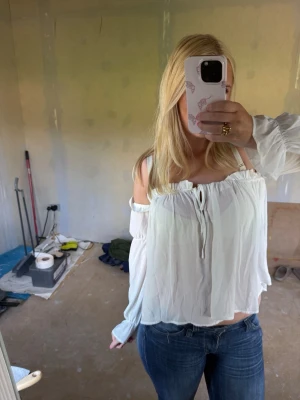 Vit offshoulder blus från NLY Trend - Supersöt vit offshoulder blus från NLY Trend med långa ärmar och volangdetaljer upptill. Blusen har en lös passform och knytning framtill vid bysten. Perfekt för en trendig och avslappnad look.
