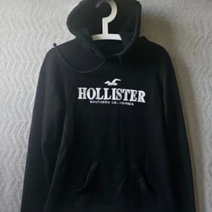 Svart hoodie från Hollister - Svart hoodie från Hollister med vit logga och text på bröstet. Klassisk känguruficka framtill och justerbar huva med snören. Tillverkad i mjukt material som passar perfekt för en avslappnad stil.
