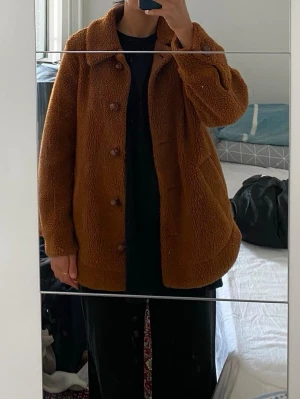 Brun teddyjacka från H&M - Säljer en brun teddyjacka från H&M i storlek 34. Jackan har klassisk krage, knäppning med bruna knappar och två fickor framtill. Materialet är mjukt och fluffigt, perfekt för kyliga dagar. Enkel och stilren design som är lätt att matcha.
