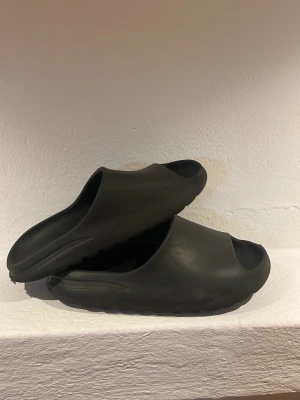 Svarta Yezzy Slides  - Säljer ett par svarta tofflor med chunky sula och öppen tå. Tillverkade i mjukt gummiliknande material för maximal komfort. Perfekta för att glida runt hemma eller på stranden. Enkel och modern design som passar till det mesta. Storlek kan diskuteras