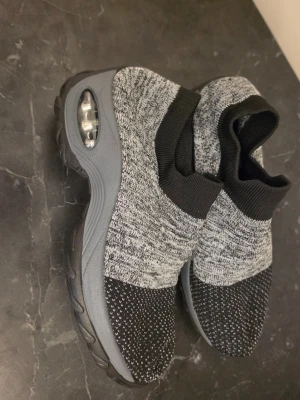 Gråsvarta stickade sneakers - Säljer ett par grå och svarta sneakers med stickad ovandel och elastisk strumpkrage. Skorna har en chunky grå sula med synlig luftbubbla i hälen och svart mönstrad tå. Perfekt för dig som gillar sportig och modern stil.