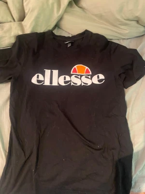 Ellesse T-shirt  - Säljer en svart t-shirt från ellesse med stor vit logga och färgglad detalj på bröstet. Klassisk passform och rund halsringning. Perfekt till jeans eller shorts för en sportig och avslappnad look.