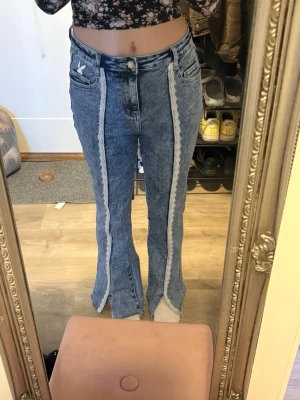 Blå utsvängda jeans med spetsdetalj - Säljer ett par blå jeans med utsvängda ben och vit spets längs framsidan. Jeansen har hög midja, klassiska fickor och en liten vit kaninlogga på fickan. Materialet är denim och modellen har slits nertill på benen.