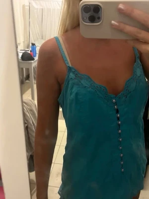 Turkos linnetopp med spetsdetaljer - Säljer en turkos linnetopp med smala axelband och spetsdetaljer längs urringningen. Toppen har dekorativa knappar framtill och är tillverkad i ett lätt material, perfekt för varma dagar.