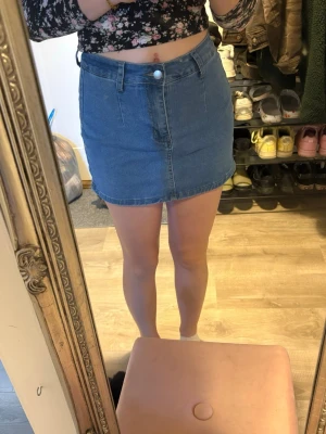 Blå jeanskjol i kort modell - Säljer en klassisk blå jeanskjol i kort modell med hög midja. Kjolen har knapp och dragkedja framtill samt fickor både fram och bak. Perfekt till sommaren och enkel att matcha med olika toppar. Har även byxor i