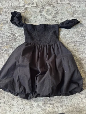 Svart kort puffklänning - Supersöt svart kort klänning med puffiga ärmar och smockad överdel. Kjolen är voluminös och ger en ballongliknande siluett. Perfekt för dig som vill sticka ut med en trendig och lekfull look.