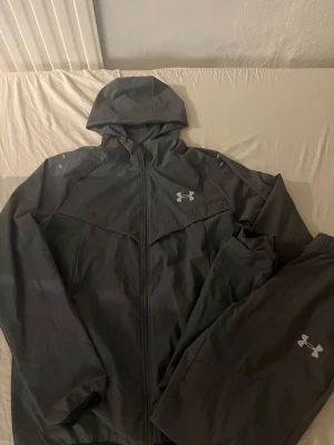 Mörk grå tracksuit från Under Armour - Snyggt träningsset från Under Armour med jacka och byxor. Jackan har huva och dragkedja, båda plaggen har logga framtill. Materialet är lätt och passar perfekt för träning eller löpning utomhus.