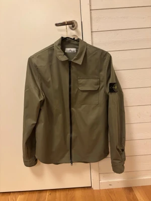 Olivgrön overshirt från Stone Island - Snygg olivgrön overshirt från Stone Island med dragkedja framtill, klassisk krage och en bröstficka med lock. Ikonisk patch på vänster ärm. Tillverkad i ett tunt, lätt material som passar perfekt för lager-på-lager. Enkel och stilren design.