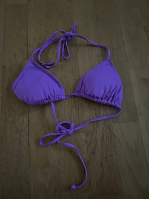 Lila trekantsbikinitopp med knyt - Snygg lila trekantsbikinitopp med knyt både i nacken och runt ryggen. Klassisk modell som är enkel att justera för bästa passform. Perfekt för lata dagar på stranden eller vid poolen.
