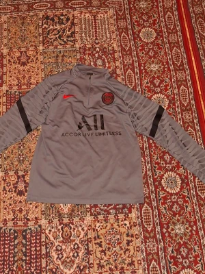 Grå PSG träningströja med dragkedja Size M PRIS KAN DISKUTERAS!! - Snygg grå långärmad träningströja från PSG med svart och rött tryck, halv dragkedja framtill och detaljerade ärmar. Nike-logga och PSG-märke på bröstet, samt texten 'Accor Live Limitless' och 'Visit Rwanda' på ryggen. Perfekt för fotbollsträning eller chill PRIS KAN DISKUTERAS Size M.