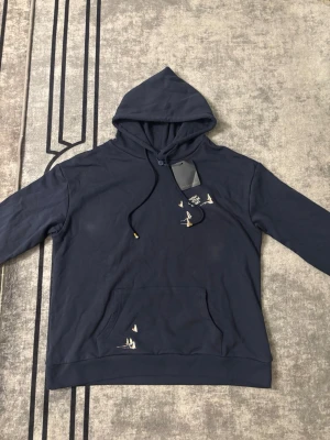 Mörkblå hoodie med fågeltryck - Snygg mörkblå hoodie med huva och dragsko. Framtill finns en känguruficka och små vita fåglar tryckta på bröstet och fickan. På ryggen syns ett stort LV-inspirerat tryck med fåglar som formar bokstäverna. Perfekt för dig som gillar streetwear och coola detaljer.