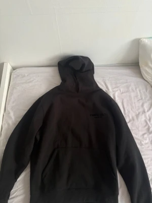 Svart Essentials Fear of God hoodie - Säljer en svart hoodie från Fear of God Essentials med stor logga på ryggen och mindre tryck på bröstet. Hoodien har en klassisk huva och stor magficka framtill. Tillverkad i mjukt bomullsmaterial som känns skönt mot huden. Pris kan diskuteras , har tags men de är inte på tröjan