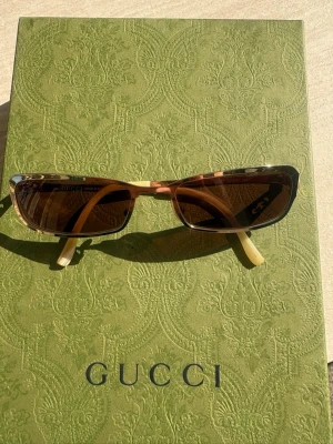 Gucci solglasögon Y2K - Snygga solglasögon från Gucci med rektangulär form och bruna bågar i transparent plast. Guldiga detaljer på sidorna och Gucci-logga på skalmen. Ljusbeige skalmtoppar ger en cool kontrast. Perfekta för dig som vill ha en trendig accessoar.