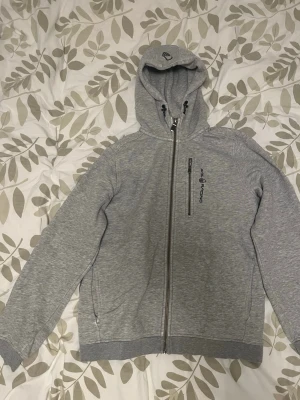 Grå sail racing hoodie - Snygg grå hoodie från Sail Racing med dragkedja hela vägen, huva med dragsko och en bröstficka med dragkedja. Tillverkad i mjukt material och har ribbade muddar vid ärmar och nederkant. Perfekt för chill dagar eller när du vill ha en sportig look.