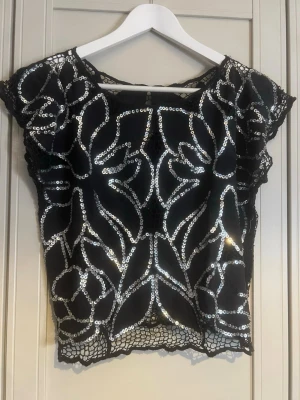 Svart blus med paljetter och spets - Säljer en svart blus med glittriga silverpaljetter i ett unikt mönster på framsidan. Blusen har korta ärmar och detaljer i spets längs kanterna. Perfekt för dig som vill sticka ut lite extra med en cool och elegant topp.