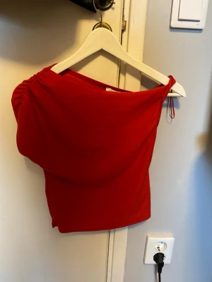 Röd topp - Snygg röd topp med rynkad design på axeln och snurrat band på andra axeln! Aldrig använd