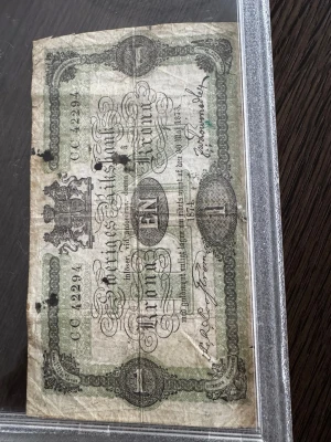 Svensk 1 krona sedel 1874 - Svensk 1 krona sedel från 1874 med detaljerat tryck, riksvapnet och text i svart och grönt. Sedeln är rektangulär och tillverkad av papper, perfekt för dig som gillar samlarprylar eller vill ha en unik historisk detalj.