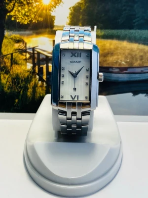 Silverfärgad rektangulär GANT damklocka - Stilren klocka från GANT med rektangulärt silverfärgat boett och matchande länkarmband i metall. Urtavlan är ljus med romerska siffror vid 12 och 6 samt små stenar som timmarkeringar. Datumvisare vid klockan 6.  Perfekt accessoar för dig som gillar klassisk och modern stil. Nytt batteri, säljes i befintligt skick enligt bilderna!