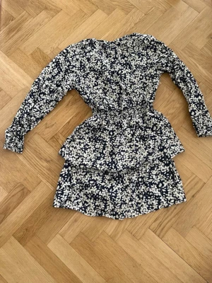 Blommig kortklänning från Lindex - Supersöt blommig kortklänning från Lindex i vitt och marinblått. Klänningen har lång ärm, markerad midja med resår och volangdetaljer på kjolen. Perfekt för sommarens alla tillfällen och gjord i mjuk bomull.