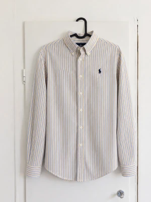 Polo Ralph Lauren randig skjorta - Klassisk långärmad skjorta från Polo Ralph Lauren med smala blå och gula ränder på vit botten. Mörkblå broderad Polo-logga på bröstet och button-down-krage. Snygg färgkombination och lätt att matcha med både jeans och chinos. 📏 Storlek: L (14-16) ✨ Skick: Mycket bra utan defekter! Hör av dig vid frågor eller om du vill ha fler bilder/mått! 📦