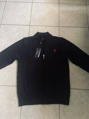 Svart stickad Ralph lauren zip  - Mörkgrå stickad kofta med hel dragkedja framtill och ribbade muddar vid ärmslut och nederkant. Liten röd broderad logga på bröstet ger en snygg detalj. Koftan har hög krage och är tillverkad i mjukt material, perfekt för lager på lager.