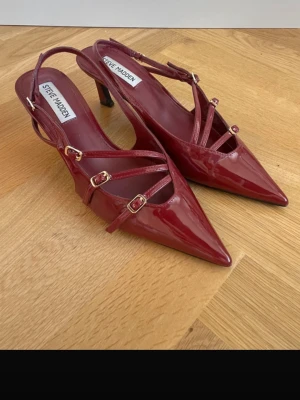 Vinröda lackpumps från Steve Madden - Snygga vinröda pumps i lack från Steve Madden med spetsig tå och smala remmar med guldfärgade spännen. Skorna har öppen häl och smal klack som ger en elegant siluett. Perfekta för dig som vill sticka ut med stilrena detaljer.