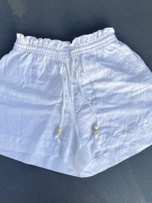 Vita linneshorts - Säljer ett par vita linneshorts med elastisk midja och snörning med pärldetaljer. Loose passform och materialet är av luftig linneblandning och är perfekta för varma dagar. I mycket bra skick endast använda fåtal gånger Två små fläckar på baksidan av shortsen men som knappt syns. (Vid önskemål kan jag tvätta de innan köp för att få bort fläckarna)