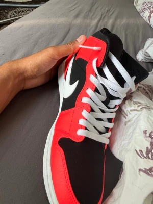 Svart och röd Nike Air Jordan 1 High - Säljer ett par Nike Air Jordan 1 High sneakers i svart och rött med vita detaljer och vit swoosh. Skorna har vita skosnören och en klassisk hög siluett. Materialet är en mix av läder och textil, och tungan har Jumpman-loggan broderad i rött.