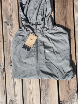 Grå vindjacka från Nike med huva - Snygg grå vindjacka från Nike med dragkedja framtill och justerbar huva. Jackan har en minimalistisk design med Nike-logga på bröstet och är tillverkad i lätt, vindtätt material. Perfekt för blåsiga dagar och enkel att packa ner i väskan.