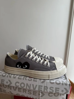 Grå Converse x CDG Play sneakers - Helt nya Grå låga Converse x Comme des Garçons Play sneakers med vit tåhätta och sula. Skorna har vita snören och det ikoniska svarta hjärta med ögon på sidan. Tillverkade i canvas med klassisk gummisula. Perfekta för en clean och stilren look.
