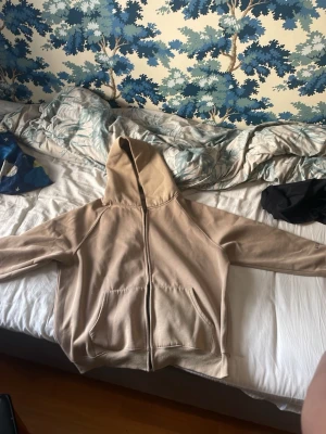 Beige Essentials hoodie med dragkedja - Säljer en beige hoodie från Essentials med dragkedja framtill och stor huva. Hoodien har två fickor på magen och Essentials-tryck på ryggen. Perfekt för dig som gillar streetwear och vill ha en enkel men snygg look.