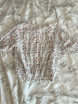 Vit blommig blus med puffärm - Söt vit blus med småblommigt mönster i rosa, blått och ljusgrönt . Blusen har puffiga korta ärmar och resår i midjan samt vid ärmsluten för en avslappnad och trendig look. Perfekt till jeans eller kjol för en fräsch sommarstil.