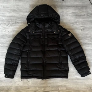 Svart pufferjacka från Bvaldi - Snygg svart pufferjacka med stor huva och två bröstfickor med lock. Jackan har quiltad design, dragkedja framtill och två sidofickor. På vänster ärm finns en liten vit detalj. Perfekt för kalla vinterdagar och riktigt varm.