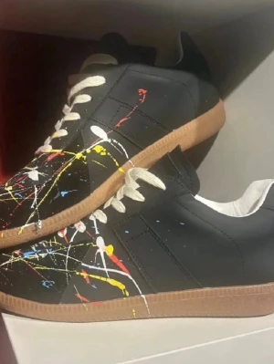Svarta sneakers med färgstänk - Svarta sneakers med vita snören och brun gummisula. Skorna har ett kreativt mönster av färgstänk i vitt, gult, rött och blått över hela ovandelen. Materialet ser ut som konstläder och modellen har lågt skaft för en avslappnad vibe.