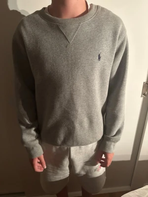 Grå sweatshirt från Ralph Lauren - Klassisk grå sweatshirt från Ralph Lauren med rund halsringning och diskret broderad logga på bröstet. Tröjan har ribbade muddar vid ärmslut och nederkant samt en relaxed passform. Perfekt till jeans eller mjukisbyxor.